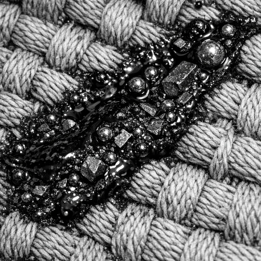 Microscopic view of 'Complex Petroleum Lipids' embedded in a cotton canvas weave. Shows the black, viscous grease trapping metallic particulates. Scientific monochrome aesthetic, sharp detail, 8k resolution.