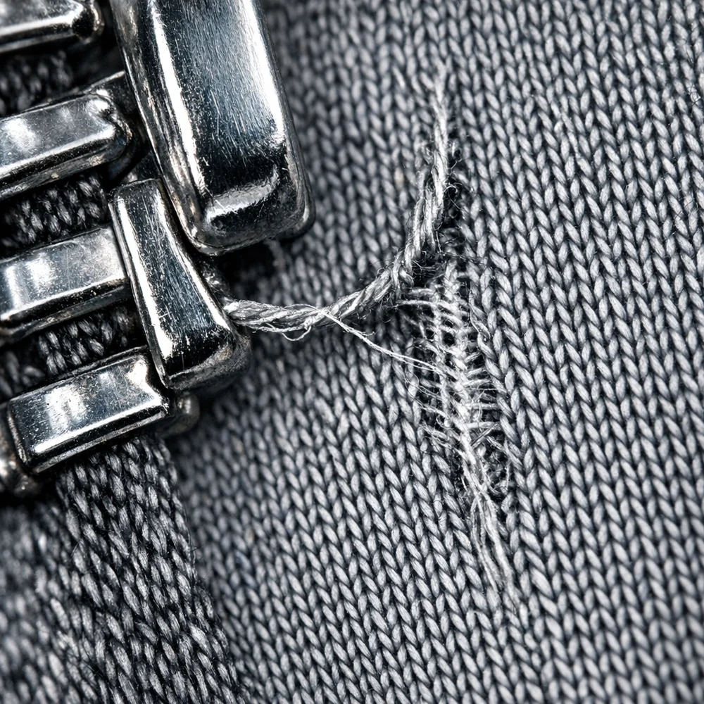 Macro shot of a metal zipper's teeth snagged onto a fine-knit synthetic fabric, causing a visible 'run' in the weave. High-contrast lighting highlights the sharp edges of the hardware. Scientific detail, 8k, clinical background.